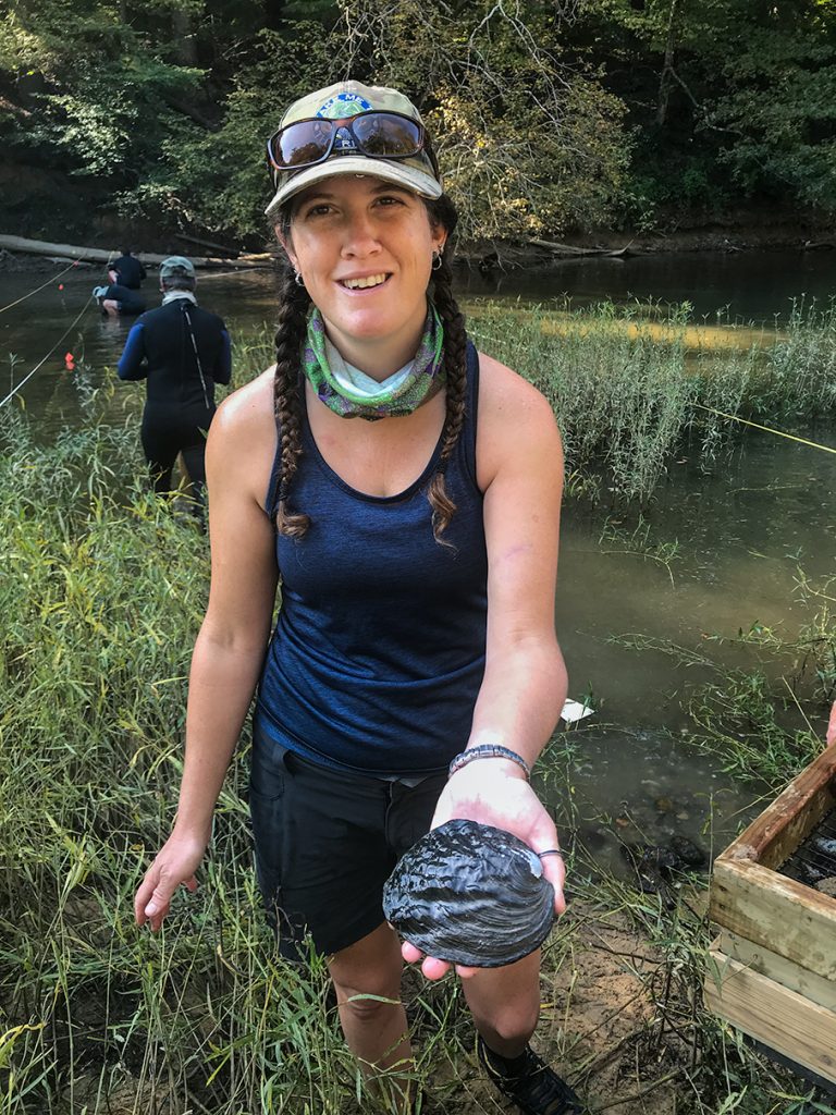 Dr. Carla Atkinson standing next to a river while holding a mussel.