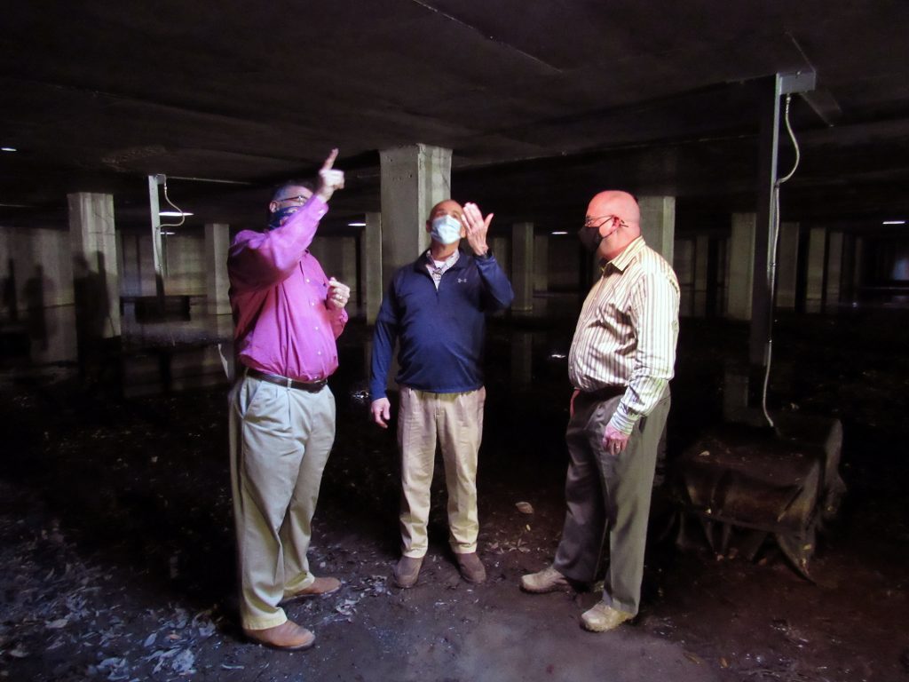Three men in a concrete room with pillars and water on the ground.