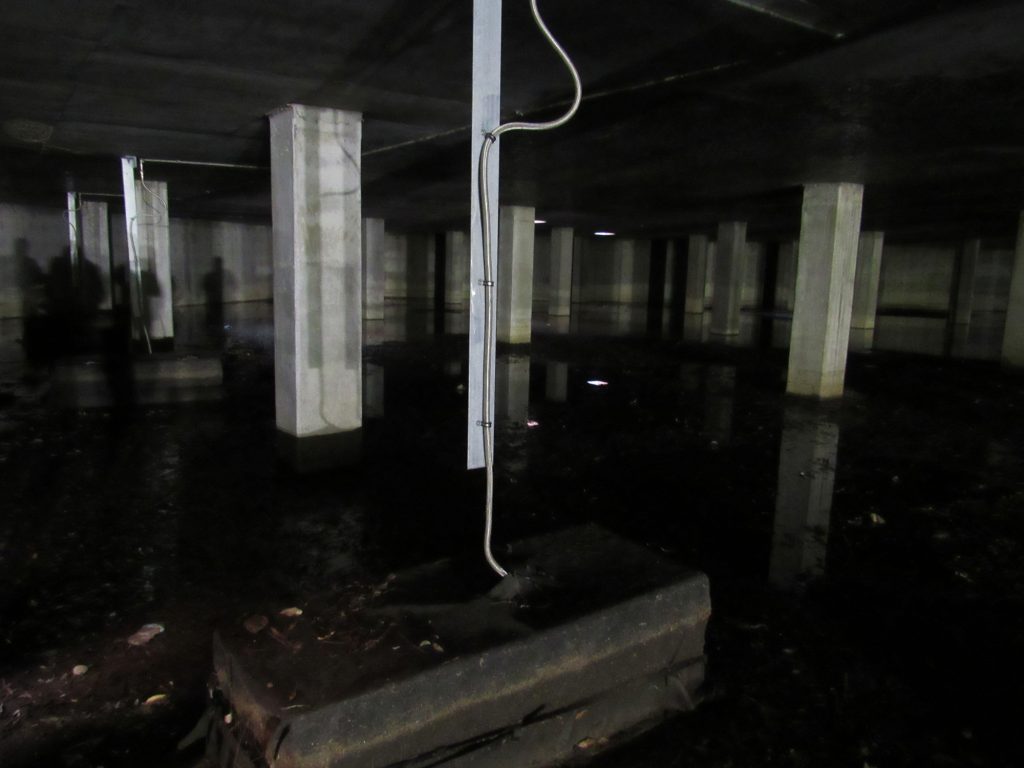 An underground concrete room with pillars and water on the ground.