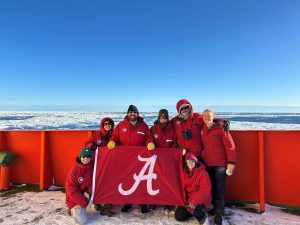 Kocot Lab team in Antarctica holding a crimson flag with The University of Alabama "A" on it in white.